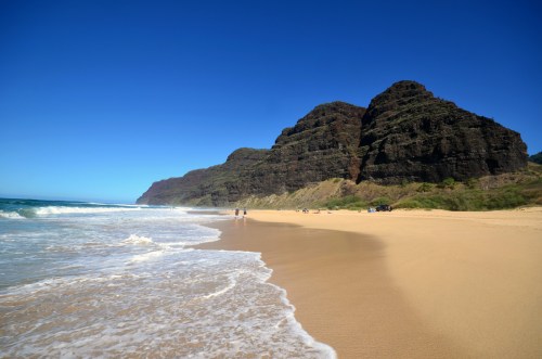 A sandy beach with gentle waves, flanked by lush green mountains under a clear blue sky.
