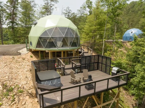 Two geodesic dome cabins on a hillside, surrounded by trees, with a deck featuring seating and a hot tub.