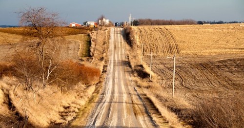 A winding dirt road stretches through a rural landscape with fields and distant farm buildings under a clear sky.