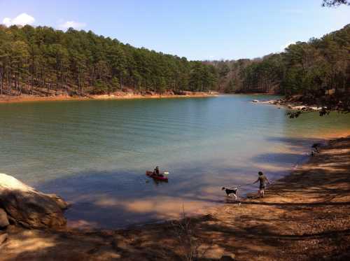 A serene lake surrounded by trees, with a person kayaking and another walking a dog along the shore.