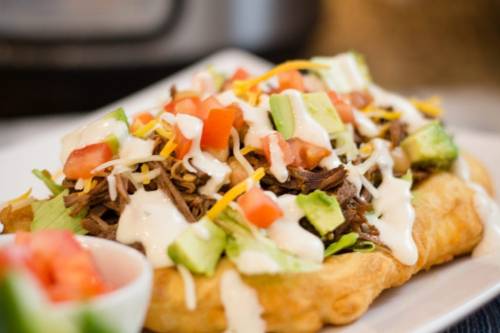 A loaded taco with shredded beef, lettuce, tomatoes, avocado, cheese, and drizzled with creamy sauce on a plate.