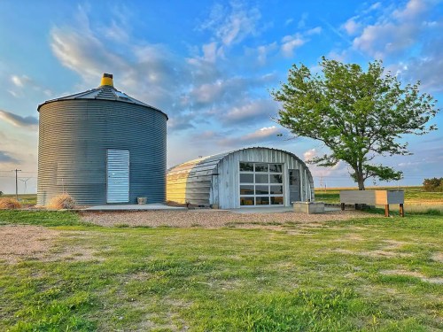 A rustic building and a silos sit on a grassy area under a blue sky with scattered clouds.