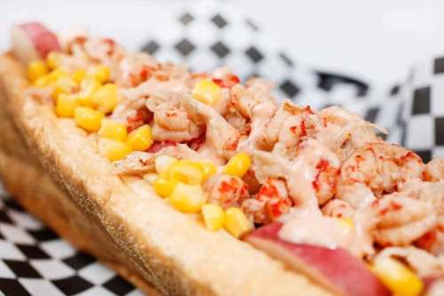 A loaded hot dog topped with shrimp, corn, and a creamy sauce, served in a toasted bun on a checkered paper background.