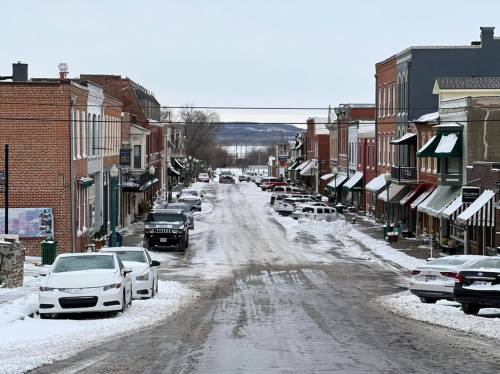 Snow-covered street lined with brick buildings and parked cars, leading to a view of a distant body of water.