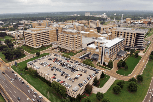 Aerial view of a large hospital complex with multiple buildings and a parking lot, surrounded by green spaces.