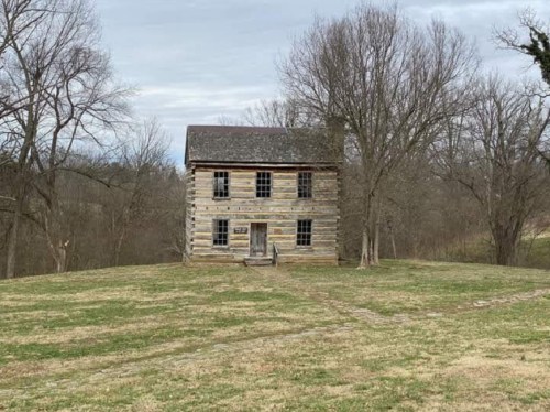 A weathered log cabin stands alone on a grassy field, surrounded by bare trees under a cloudy sky.
