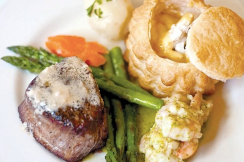 A gourmet plate featuring filet mignon, shrimp, asparagus, carrots, and a pastry filled with creamy sauce.