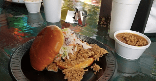 A pulled pork sandwich with coleslaw on a black plate, alongside a cup of pulled pork and two white cups.