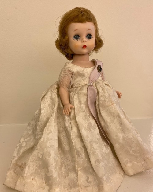 A vintage doll with curly hair, wearing a cream-colored lace dress with a pink accent and a small button.