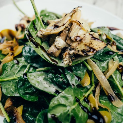 A fresh salad with spinach, artichokes, and drizzled balsamic glaze on a white plate.