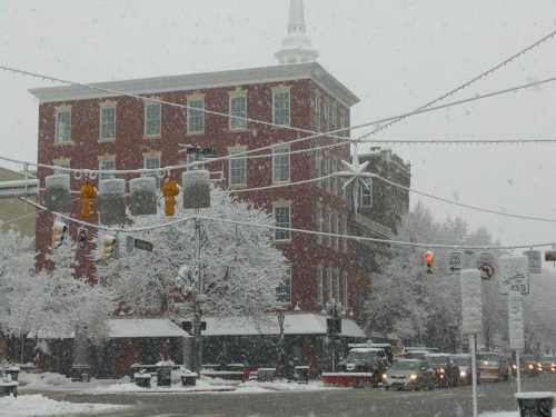 A snowy street scene featuring a red brick building, traffic lights, and trees covered in white snowflakes.