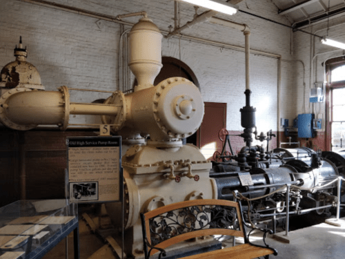 Historic pump room featuring large machinery and a display sign, set in a brick-walled industrial space.