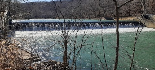 A serene view of a dam with water cascading over it, surrounded by trees and a clear blue sky.