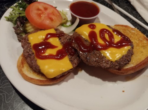 A plate with a cheeseburger featuring two patties, melted cheese, ketchup, and a side of salad and dipping sauce.