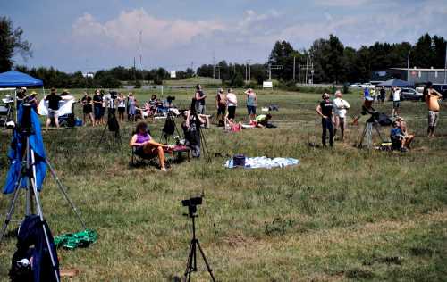 A crowd of people stands and sits in a grassy field, some with cameras, under a clear blue sky.