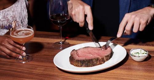A person slices into a medium-rare roast beef on a plate, with a cocktail and wine glass nearby.