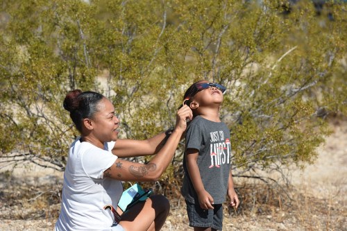 A woman adjusts sunglasses on a young boy, both smiling, with desert vegetation in the background.