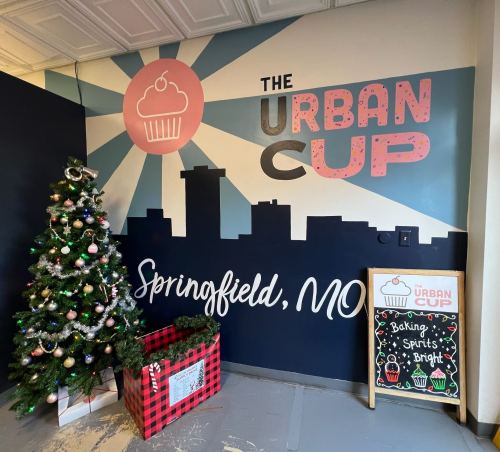 A festive corner featuring a decorated Christmas tree, a gift box, and a mural for "The Urban Cup" in Springfield, MO.