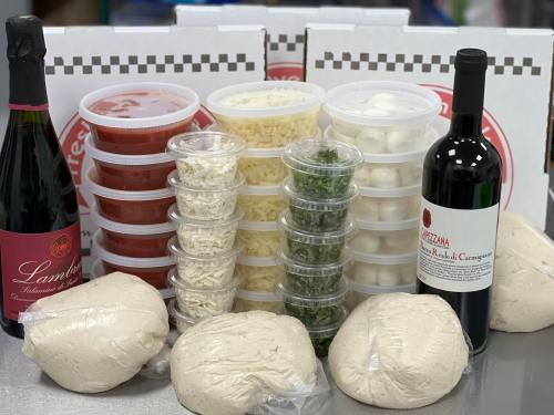 A variety of pizza ingredients including dough, sauces, cheeses, herbs, and wine, arranged with pizza boxes in the background.