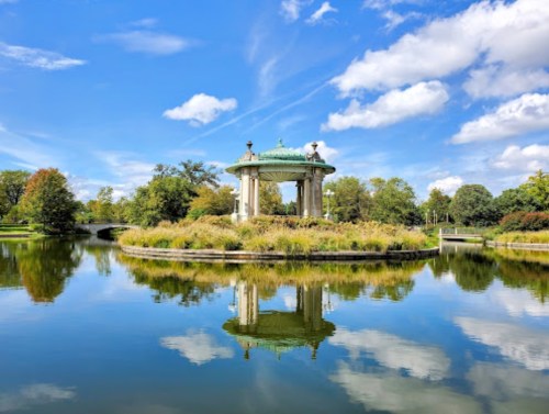 A serene park scene featuring a gazebo on an island, surrounded by water and lush greenery under a blue sky.