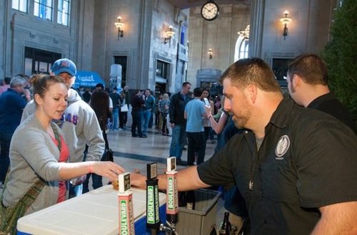 A bartender serves a drink to a customer at a busy event in a spacious venue with people mingling in the background.