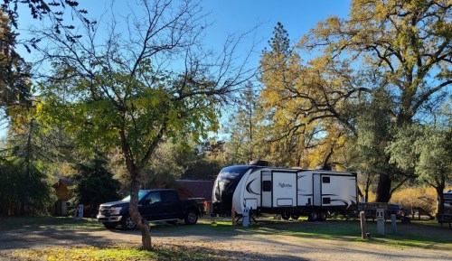 A camper trailer parked beside a black truck in a scenic campsite with autumn trees and clear blue skies.
