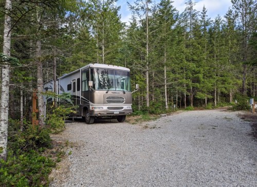 A silver RV parked on a gravel road surrounded by tall green trees in a forested area.