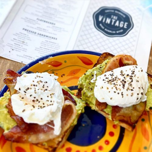 A colorful plate with two slices of toast topped with avocado, bacon, and poached eggs, alongside a menu.