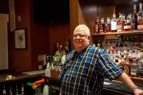 A smiling man in a plaid shirt stands at a bar with shelves of various liquor bottles in the background.
