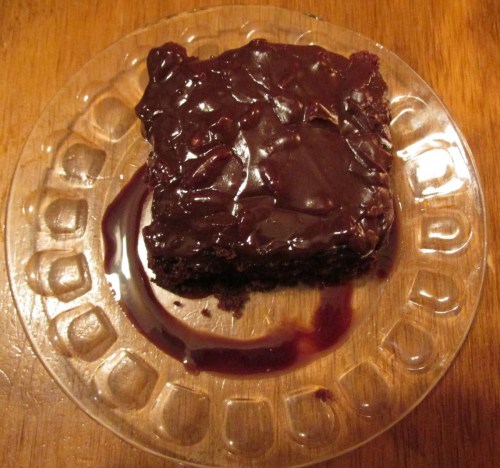 A slice of chocolate cake with glossy frosting on a clear plate, resting on a wooden surface.