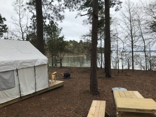 A tent by a lake surrounded by trees, with wooden picnic tables and a fire pit nearby.