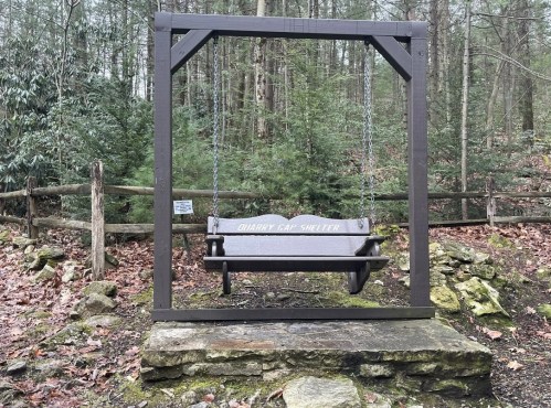 A wooden swing labeled "Quarry Gap Shelter" surrounded by trees and a stone base in a forested area.