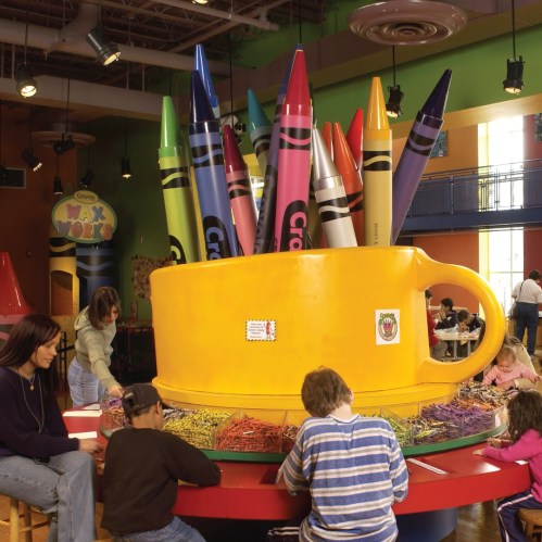 A colorful play area with large crayons and children engaged in art activities around a giant yellow cup.