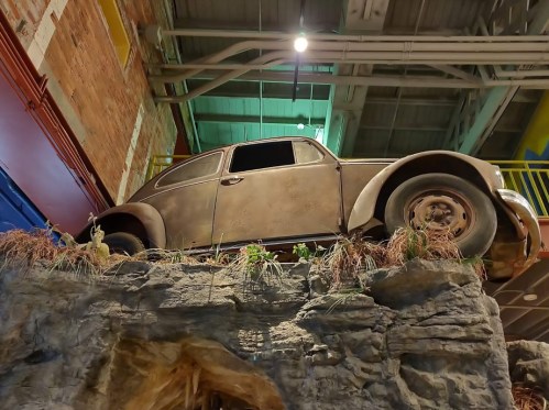 A vintage car perched on a rocky ledge, surrounded by greenery, inside a building with industrial decor.