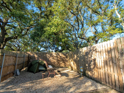 A tent set up in a sandy area surrounded by tall trees and a wooden fence under a clear blue sky.
