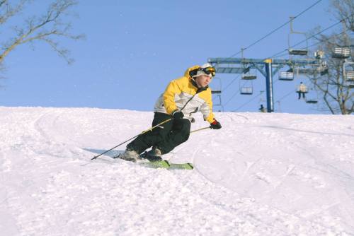 A skier in a yellow and white jacket navigates a snowy slope under a clear blue sky, with a ski lift in the background.