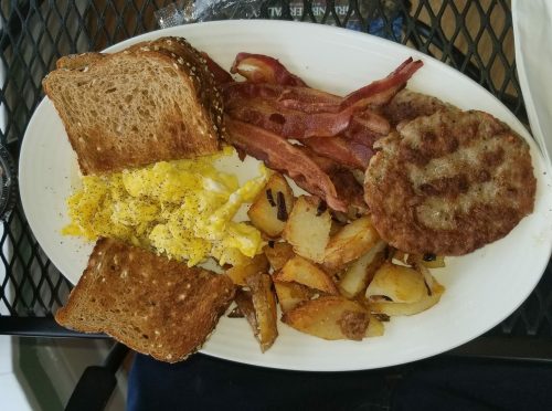 A plate of breakfast featuring scrambled eggs, bacon, sausage patty, roasted potatoes, and two slices of whole grain toast.