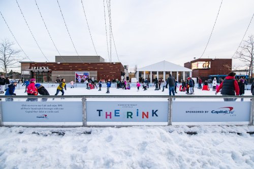 A busy ice skating rink with families and children enjoying the winter day, surrounded by snow and buildings.