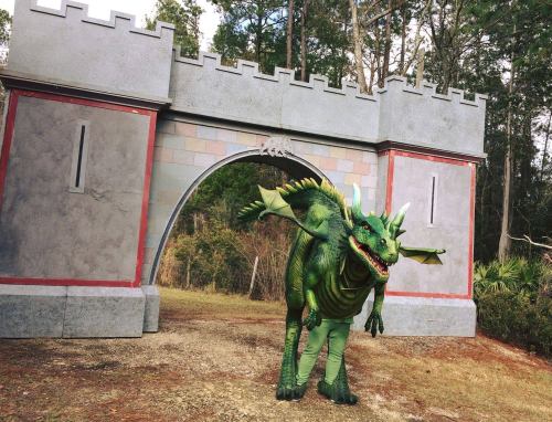 A person in a green dragon costume stands in front of a castle-like structure in a wooded area.
