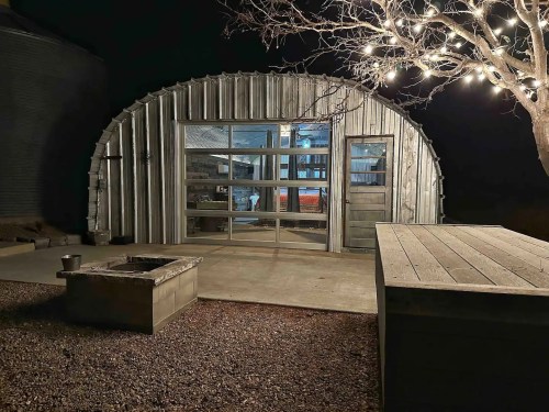 A metal arch-shaped building at night, with a lit tree nearby and a fire pit in the foreground.