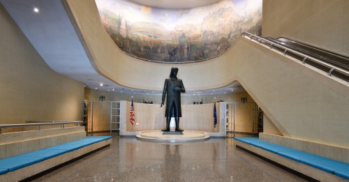 A spacious hall featuring a statue of a man, surrounded by murals and blue benches, with flags in the background.