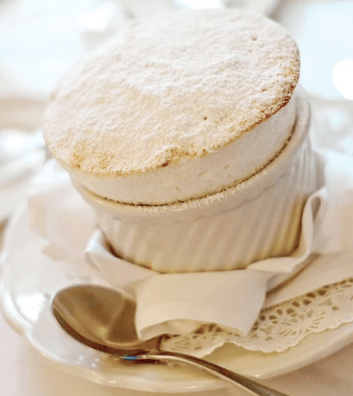 A fluffy soufflé dusted with powdered sugar, served in a white ramekin on a plate with a spoon.