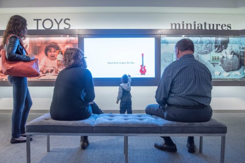 A child stands in front of a screen in a museum, while three adults sit on a bench, engaged with the exhibit.