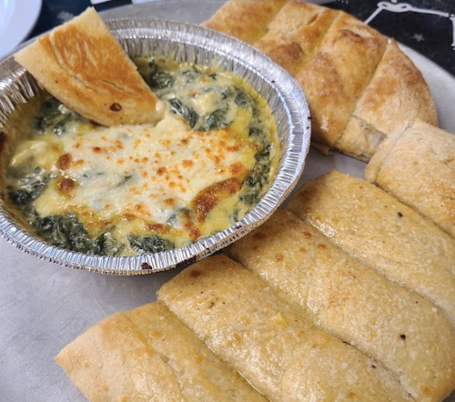 A dish of cheesy spinach dip in a foil container, with breadsticks for dipping, arranged on a silver platter.