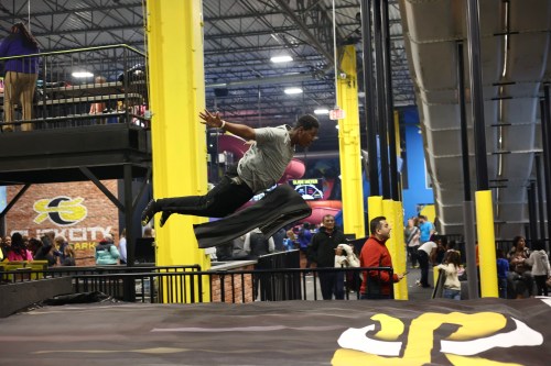 A person jumps mid-air at a trampoline park, with spectators in the background enjoying the lively atmosphere.