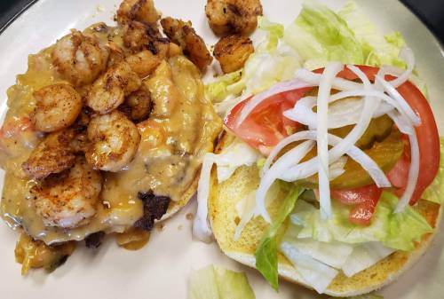 A plate with seasoned shrimp over a savory sauce, alongside a bun topped with lettuce, tomato, onions, and pickles.