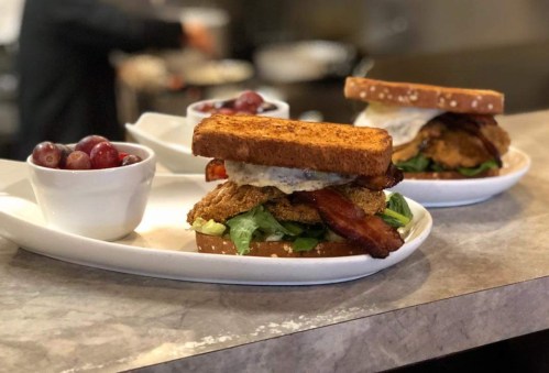 A hearty sandwich with fried chicken, bacon, and lettuce on toasted bread, served with a side of grapes.