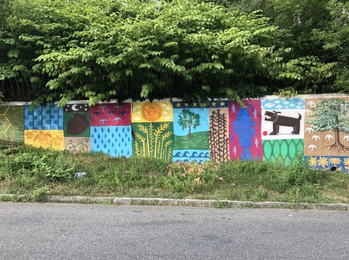 Colorful mural depicting nature scenes, including trees, water, and animals, on a wall surrounded by greenery.