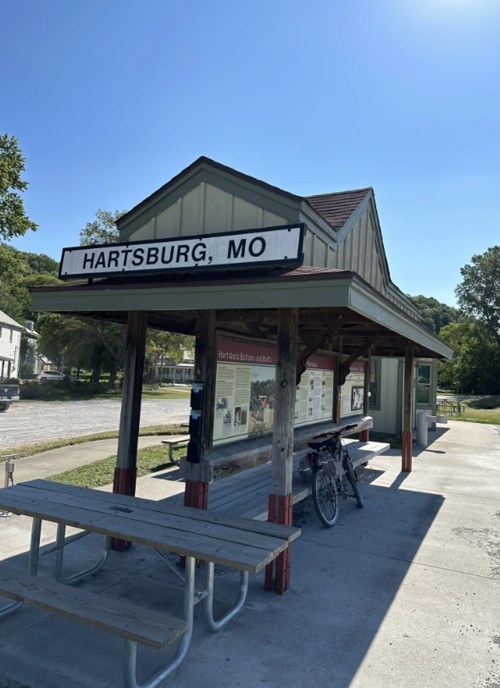 A small, rustic building with a sign reading "Hartsburg, MO," featuring picnic tables and a bicycle nearby.