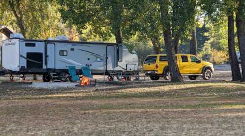 A camper trailer parked near a yellow truck, with a fire pit and chairs set up under trees in a grassy area.